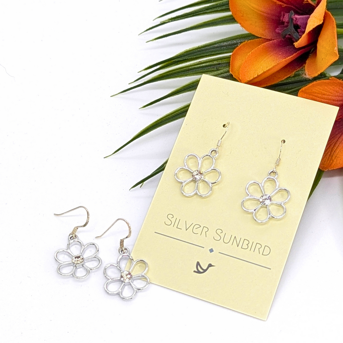 Sparkly Silver Daisy Earrings from Silver Sunbird are shown on a branded card with one pair on display and another beside it. An orange lily and green leaves in the background highlight these floral earrings.