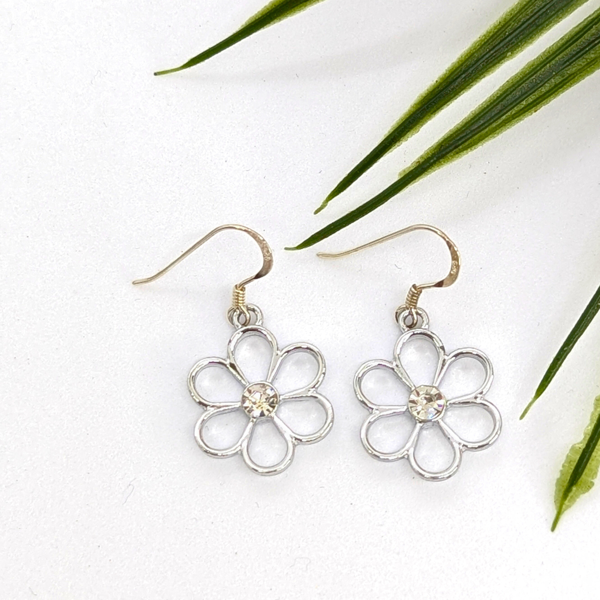 The Sparkly Silver Daisy Earrings by Silver Sunbird feature clear gem centers and are shown on a white surface beside green leaves.