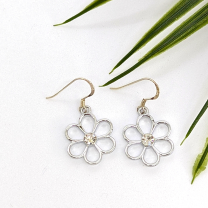 The Sparkly Silver Daisy Earrings by Silver Sunbird feature clear gem centers and are shown on a white surface beside green leaves.