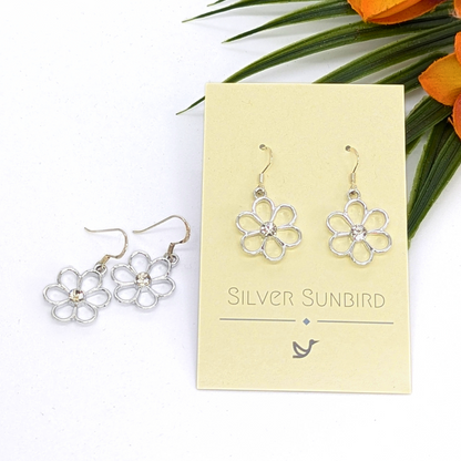 Sparkly Silver Daisy Earrings by Silver Sunbird are shown on a branded card, with one pair on the card and another beside it. Orange flowers and green leaves create a vibrant backdrop for these floral earrings.