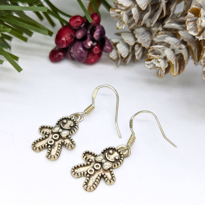 The Jolly Gingerbread Men Earrings by Silver Sunbird rest on a white surface beside pine cones, green pine needles, and red berries, adding a cheerful holiday touch.