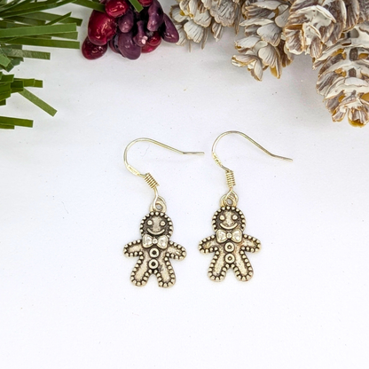 The Jolly Gingerbread Men Earrings by Silver Sunbird are crafted from 925 sterling silver with festive details, shown on a white surface with snowy pinecones, pine needles, and berries—ideal holiday jewelry.