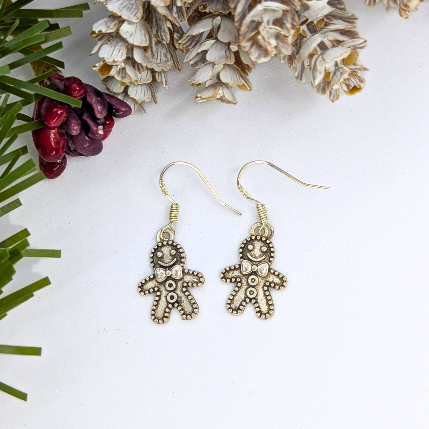 The Silver Sunbird Jolly Gingerbread Men Earrings, crafted from 925 sterling silver, are showcased on a white surface with pinecones, festive pine needles, and red berries—making them ideal holiday jewelry.