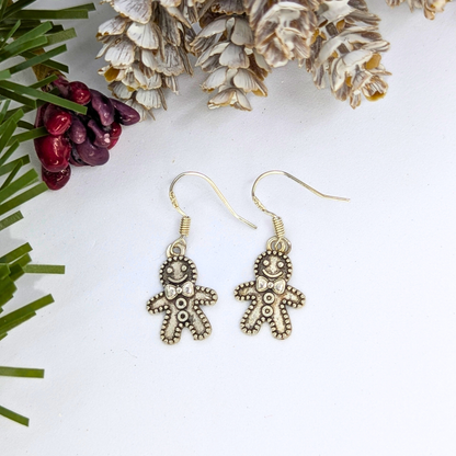 The Silver Sunbird Jolly Gingerbread Men Earrings, crafted from 925 sterling silver, are showcased on a white surface with pinecones, festive pine needles, and red berries—making them ideal holiday jewelry.