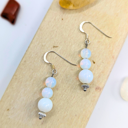 Opal Dangle Earrings by Silver Sunbird feature handmade designs with stacked opal-like iridescent beads in a subtle color range, silver hooks, and ornate bead caps on a light wood backdrop.
