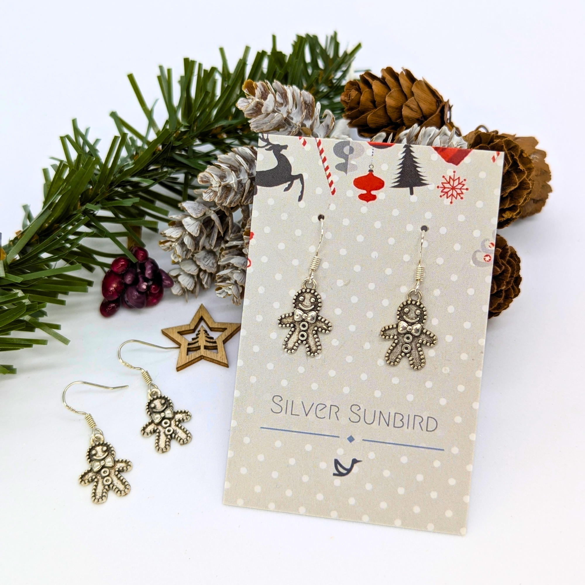 The Jolly Gingerbread Men Earrings by Silver Sunbird, crafted from 925 sterling silver, are featured on a festive polka dot card amid pinecones and evergreens—a perfect holiday jewelry gift.