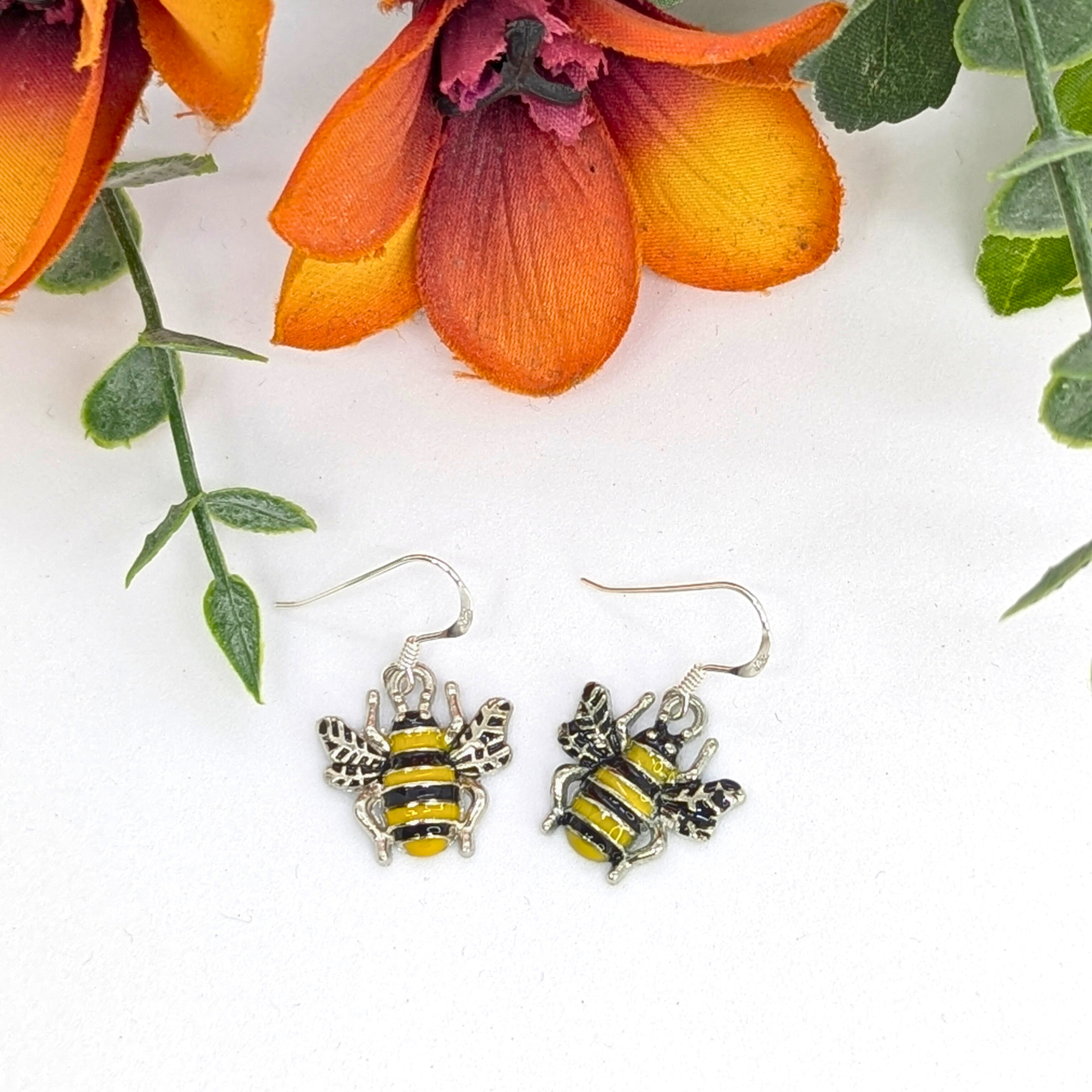 The Silver Sunbird Bumble Bee Earrings, featuring yellow and black stripes, rest on a white surface amidst orange flowers and green leaves.
