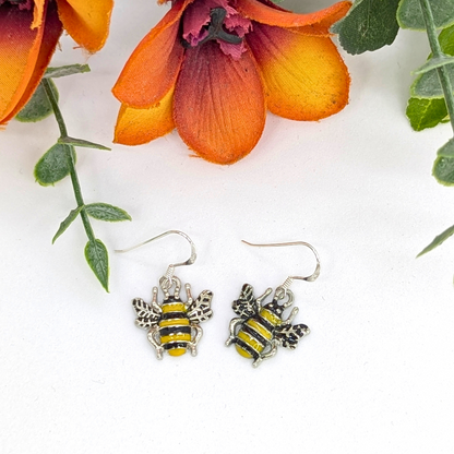 The Silver Sunbird Bumble Bee Earrings, featuring yellow and black stripes, rest on a white surface amidst orange flowers and green leaves.