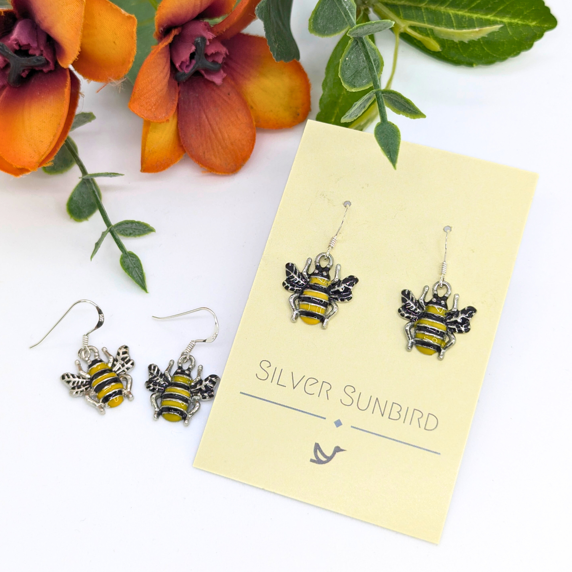 Bumble Bee Earrings by Silver Sunbird, featuring yellow and black enamel stripes, are shown on a branded card beside a matching pair. The display is set against a white background with orange flowers and green leaves.