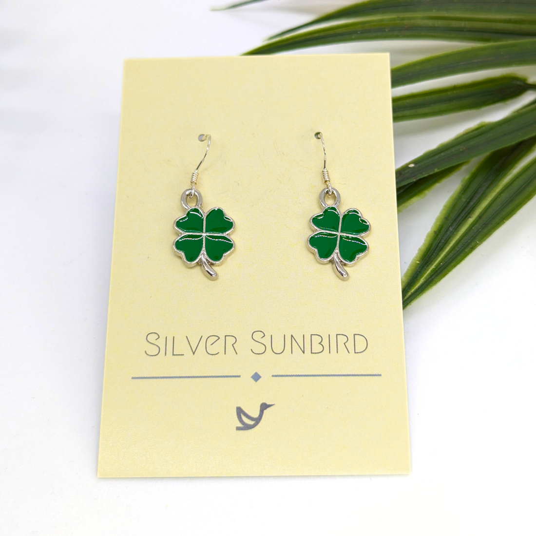 A pair of Lucky Four Leaf Clover Earrings by Silver Sunbird is displayed on a branded card with green leaves in the background. These handmade earrings bring a stylish, lucky accent to any outfit.