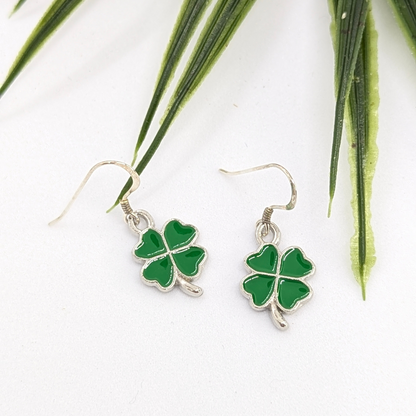 The Silver Sunbird Lucky Four Leaf Clover Earrings, handmade from sterling silver with green enamel clover charms, are displayed on a white surface beside long, slender green leaves.