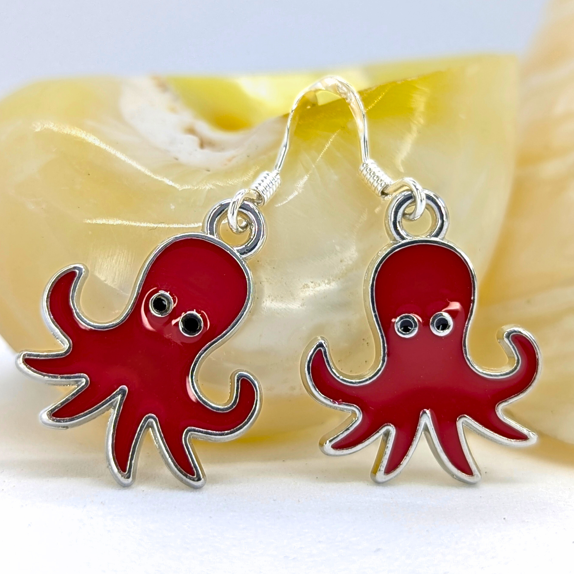 Silver Sunbird’s Red Octopus Earrings feature vivid enamel, silver outlines, and black eyes—displayed before a pale seashell. A perfect choice for ocean-inspired jewelry lovers.