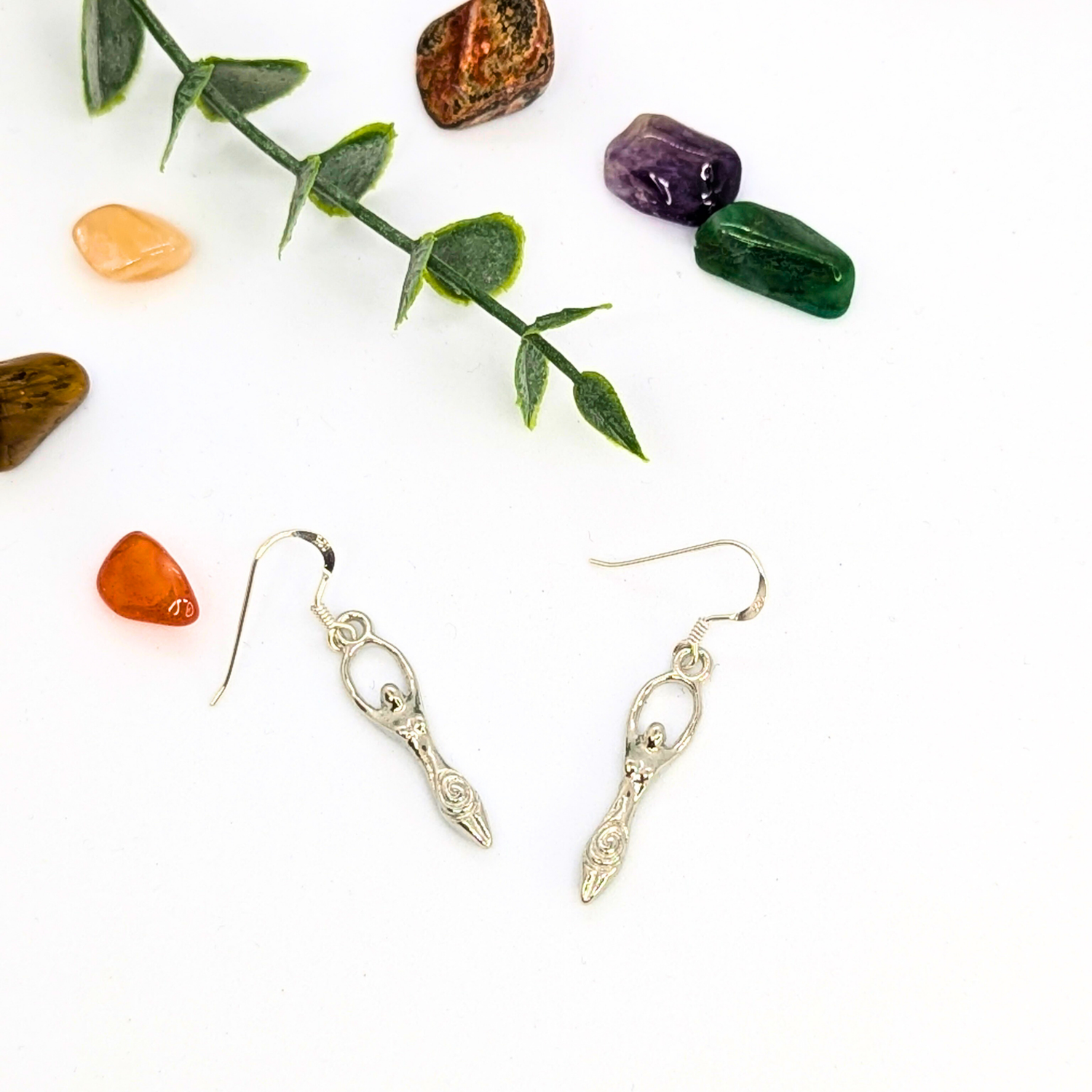 A pair of Silver Sunbird Fertility Goddess Dangle Earrings rests on a white surface beside a green leafy twig and colorful polished stones—ideal for adding boho flair with handmade goddess jewelry.
