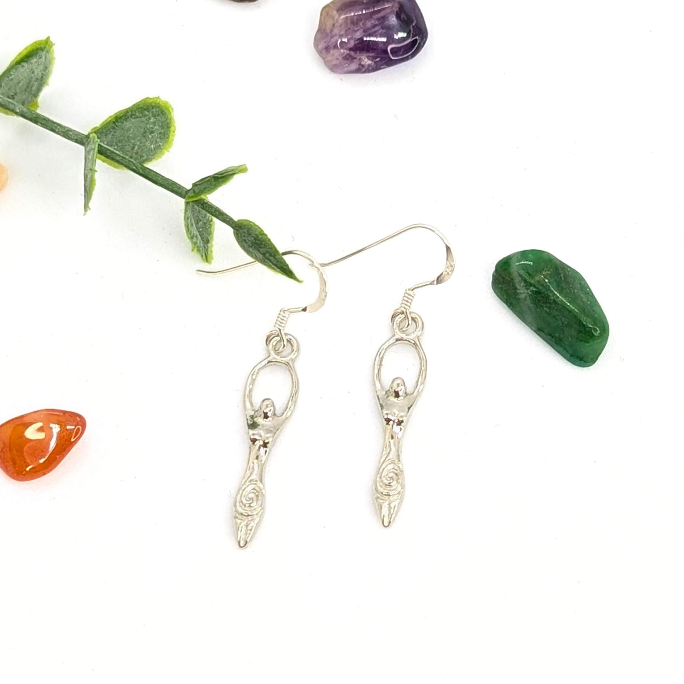 Silver Sunbird’s Fertility Goddess Dangle Earrings with raised arms are shown on a white background, accented by green leaves and colorful stones—perfect handmade goddess jewelry for a boho look.