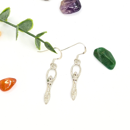 Silver Sunbird’s Fertility Goddess Dangle Earrings with raised arms are shown on a white background, accented by green leaves and colorful stones—perfect handmade goddess jewelry for a boho look.