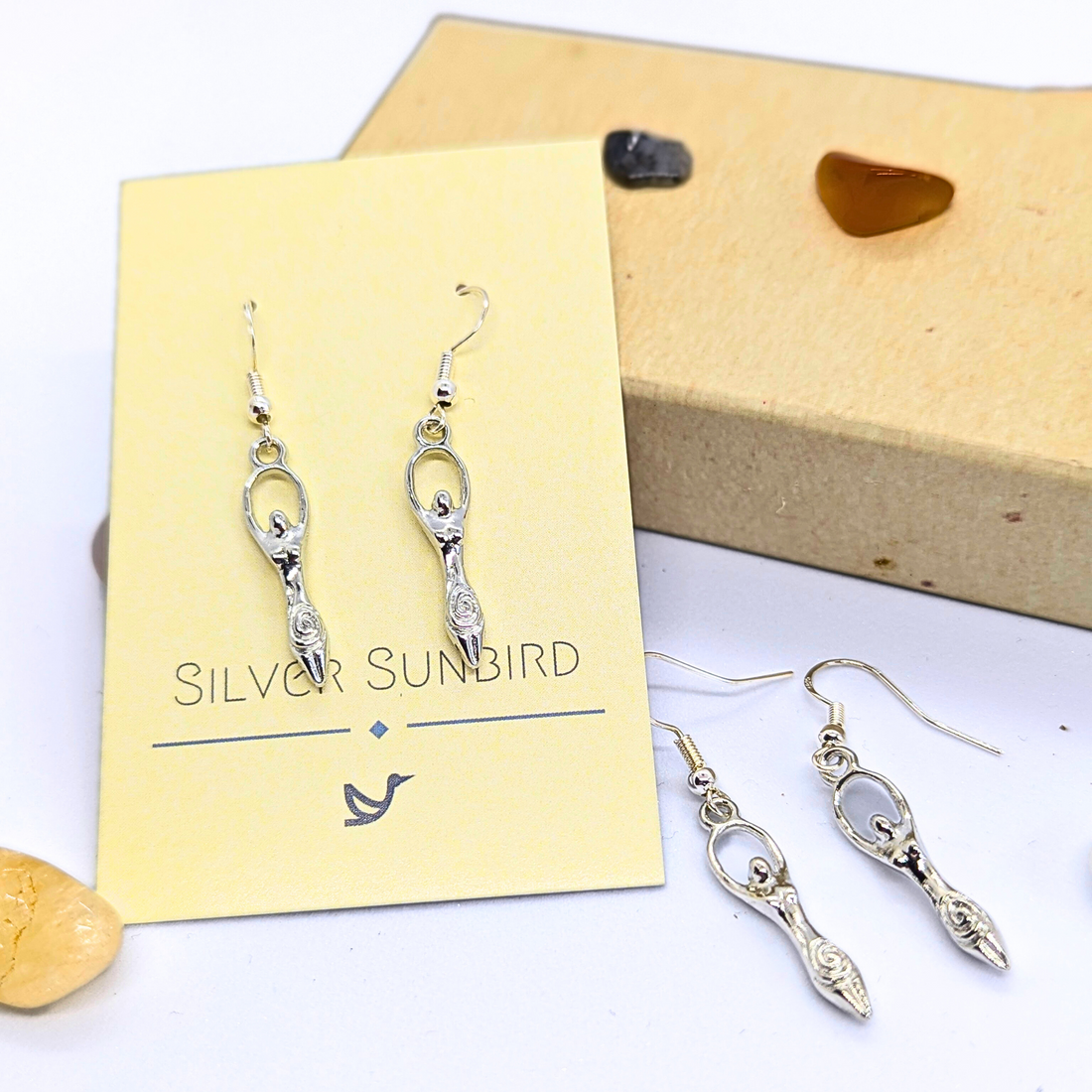 Two pairs of Silver Sunbird Fertility Goddess Dangle Earrings are displayed: one on a yellow branded card, surrounded by small stones and a brown box—perfect handmade goddess jewelry for those who love unique earrings.