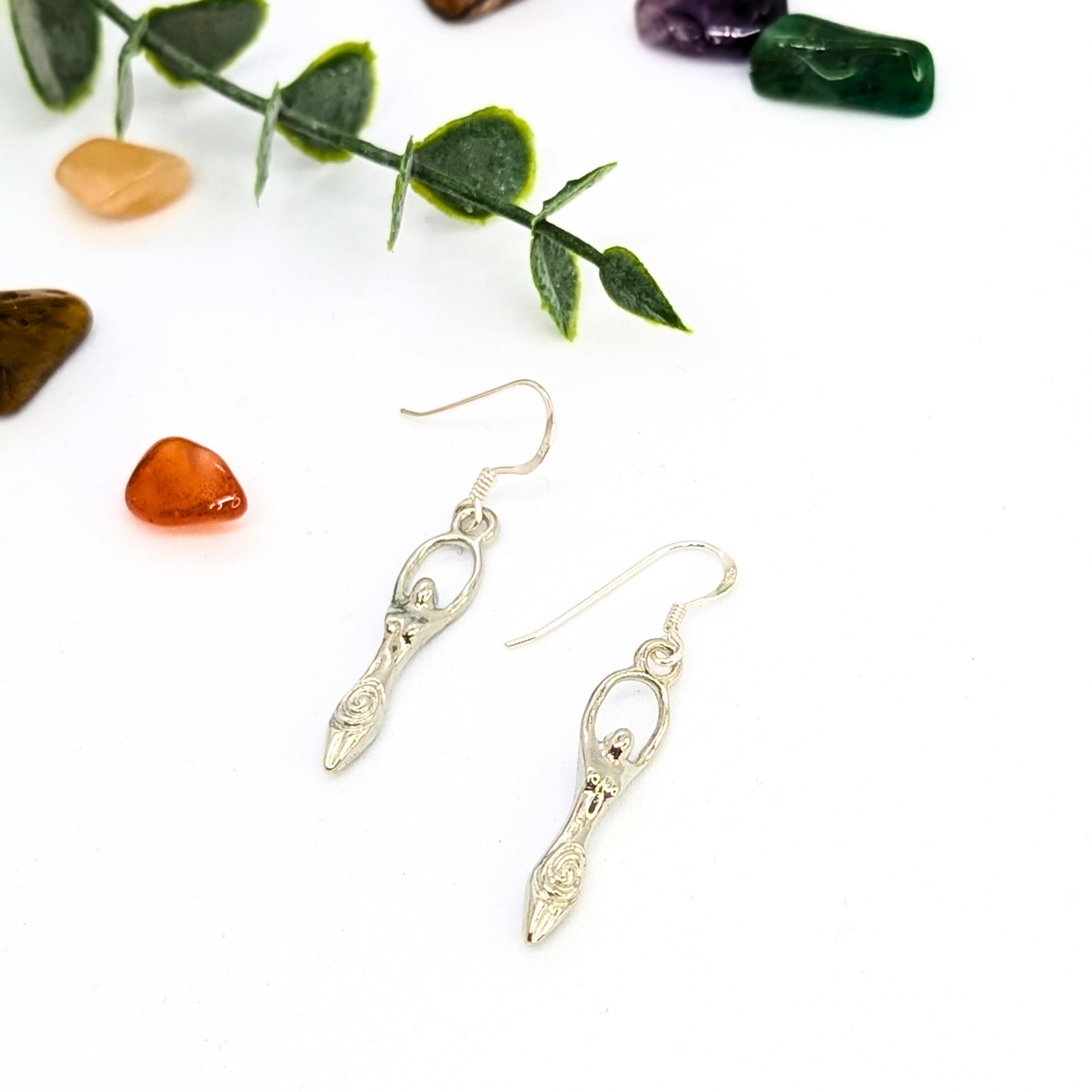 The Fertility Goddess Dangle Earrings by Silver Sunbird are shown on a white surface with green leaves and colorful stones—an ideal choice for fans of handmade goddess jewelry.