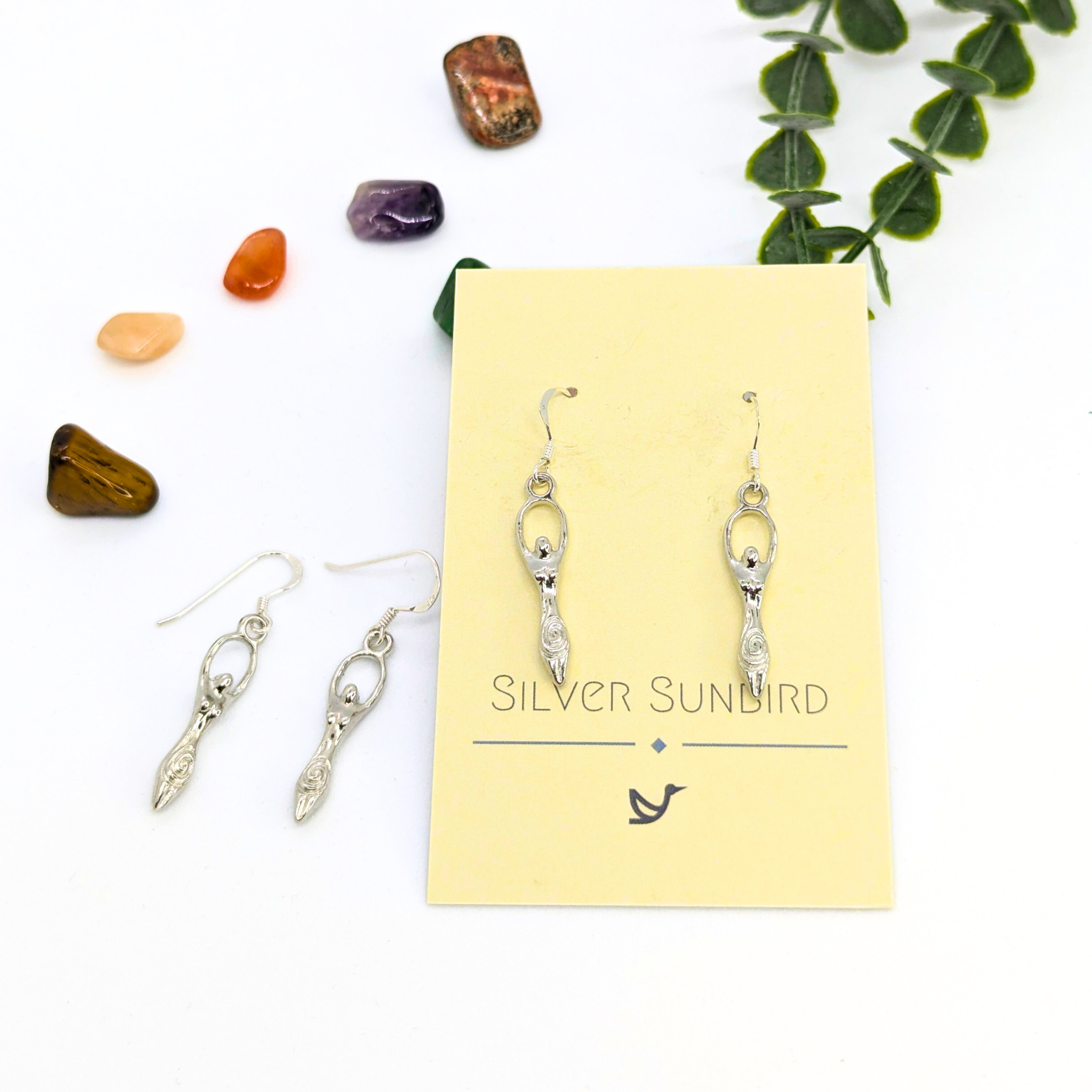 A pair of Fertility Goddess Dangle Earrings by Silver Sunbird, displayed on a branded card with polished stones and green leaves, perfect for fans of handmade goddess jewelry.