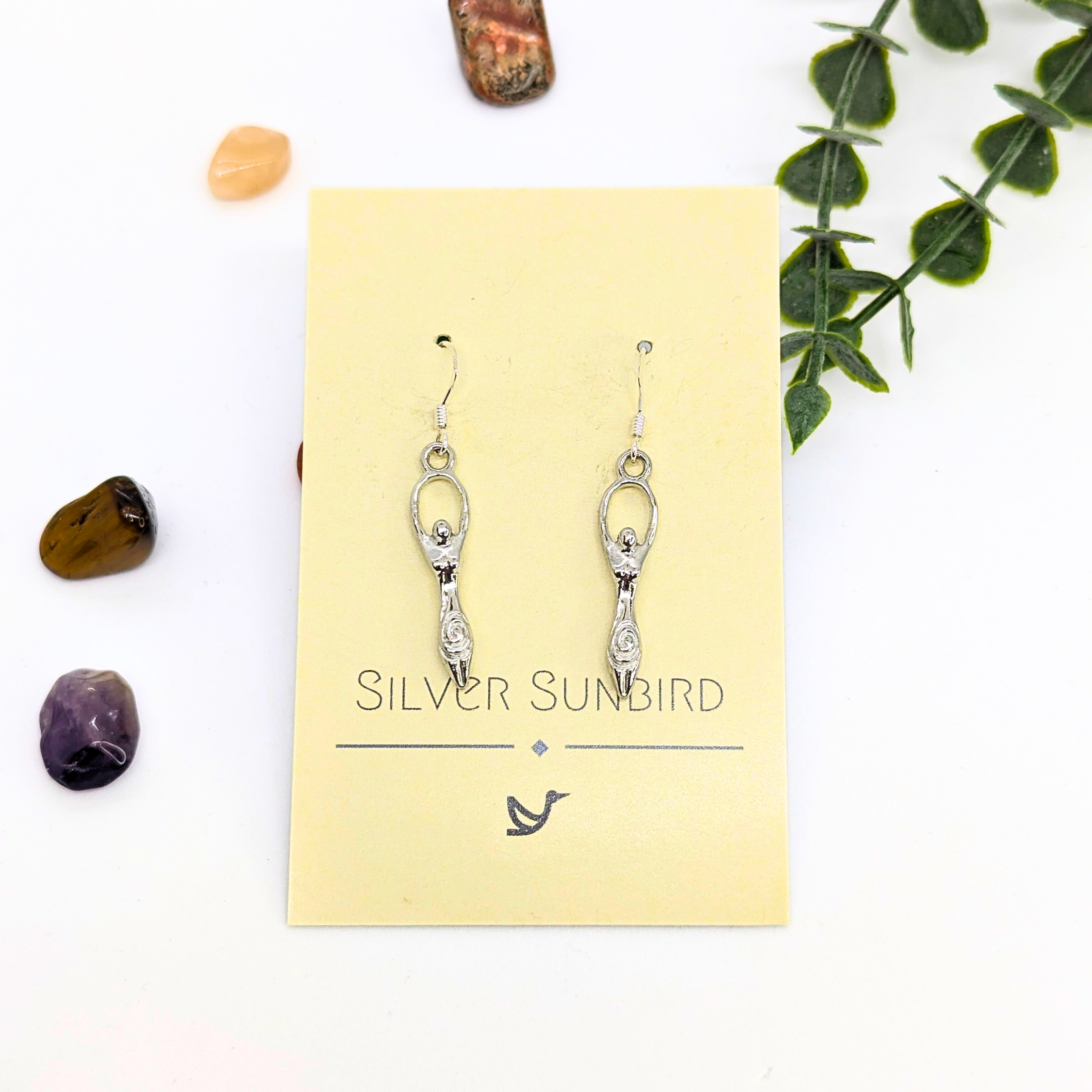 A pair of Fertility Goddess Dangle Earrings by Silver Sunbird, featuring abstract figure designs, displayed on a branded card with polished stones and greenery—a perfect handmade goddess jewelry accent.