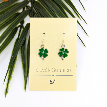 A pair of Lucky Four Leaf Clover Earrings by Silver Sunbird, featuring green enamel charms, is displayed on a branded card against a white background with green leaves in the corner.