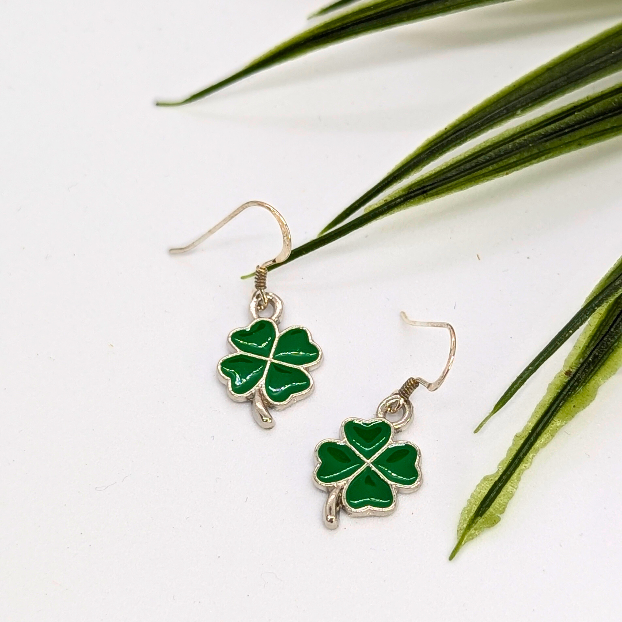 The Lucky Four Leaf Clover Earrings by Silver Sunbird are sterling silver with green enamel clover charms, shown on a white surface accented by green plant leaves.
