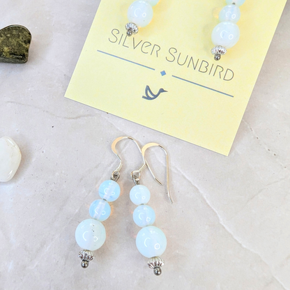 Opal Dangle Earrings by Silver Sunbird, featuring light blue beads and silver accents, are displayed on a brand card. The backdrop includes a marble surface adorned with a small dark stone and a white stone fragment.