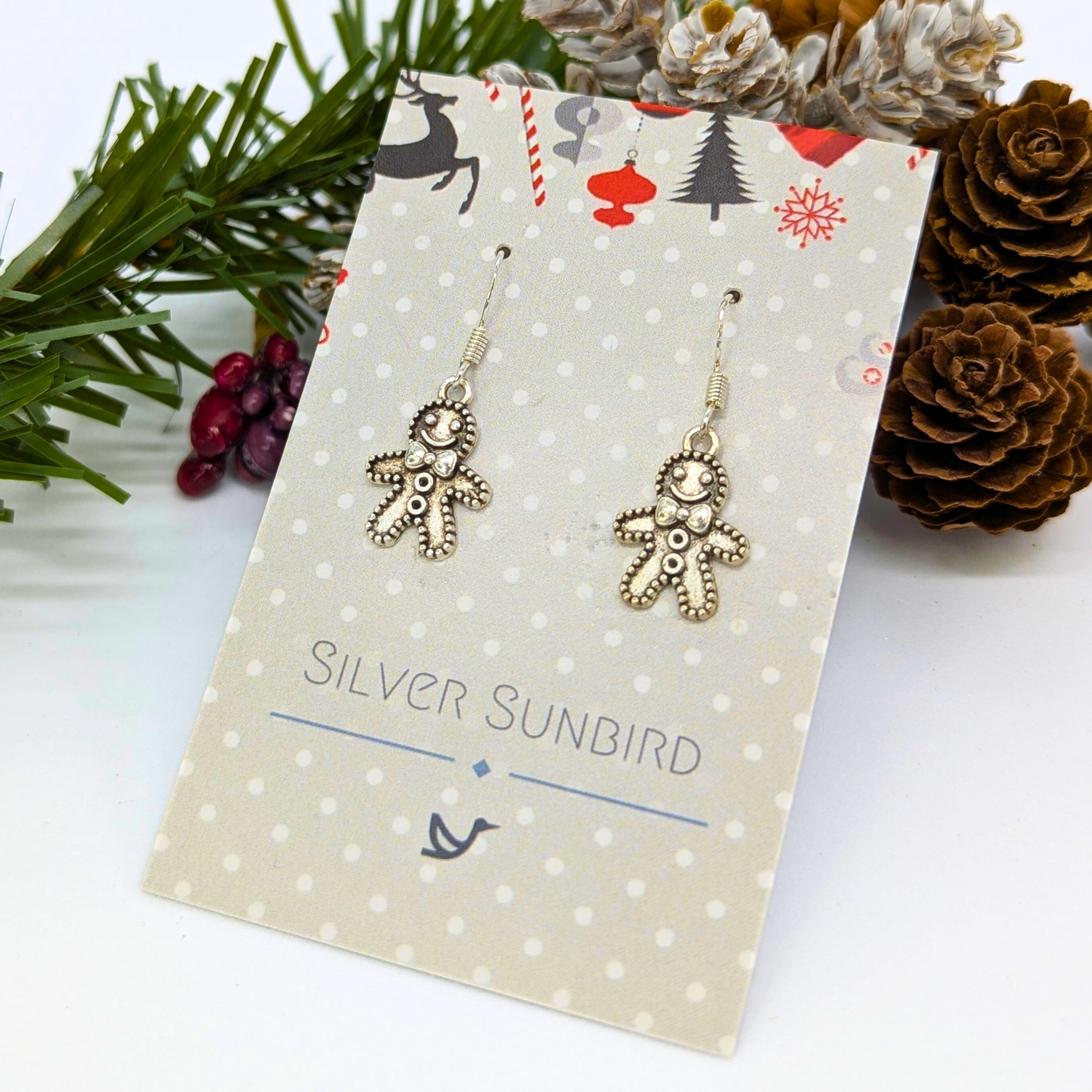 Jolly Gingerbread Men Earrings by Silver Sunbird on a polka dot card, nestled among pinecones and festive greenery—cheerful holiday jewelry perfect for the season.