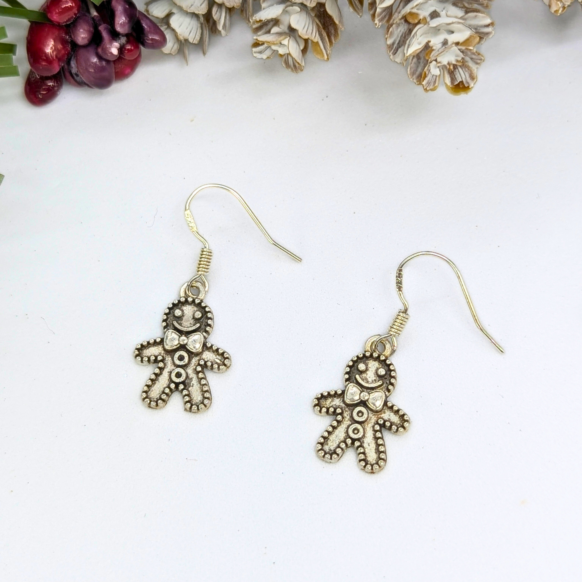 Jolly Gingerbread Men Earrings by Silver Sunbird, featuring charming handmade details, are shown on a white surface with pine cones and berries in the background.