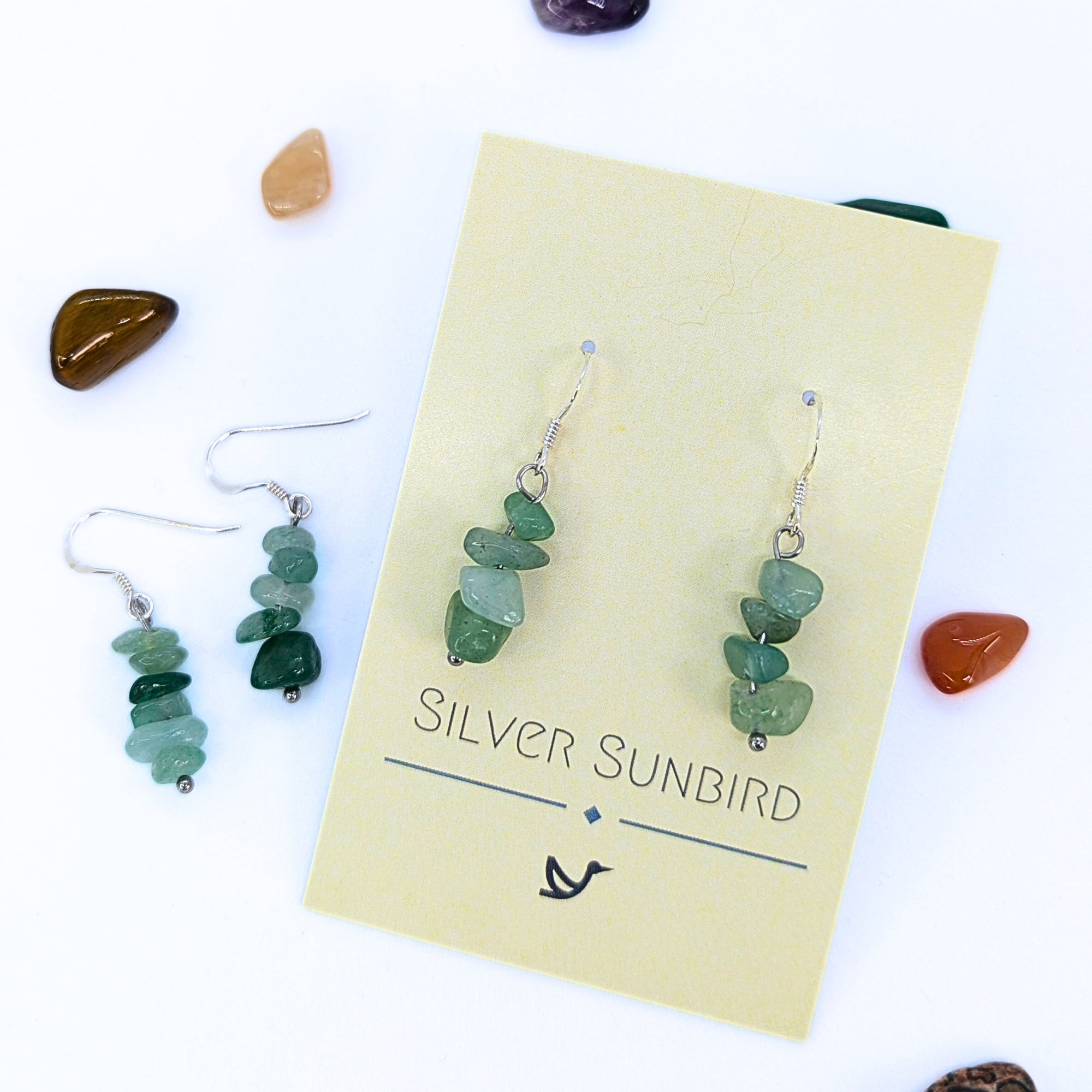 Green Aventurine Earrings by Silver Sunbird are shown on a branded card, with a matching pair and colorful polished stones nearby—a beautiful display of handmade gemstone jewelry.