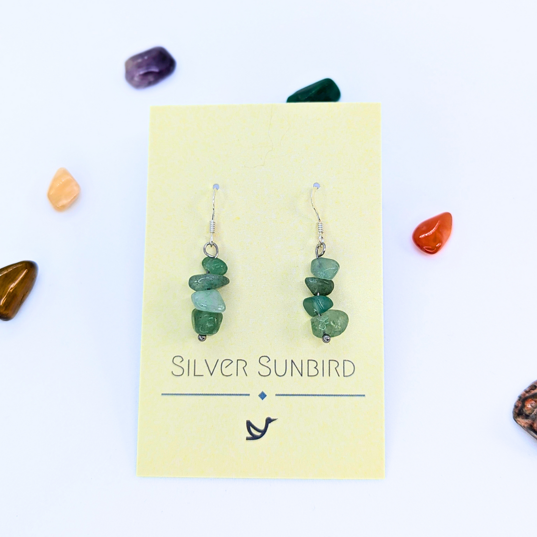 Green Aventurine Earrings by Silver Sunbird with stone chips dangle on a yellow card, surrounded by colorful tumbled stones—unique handmade gemstone jewelry displayed on a crisp white background.