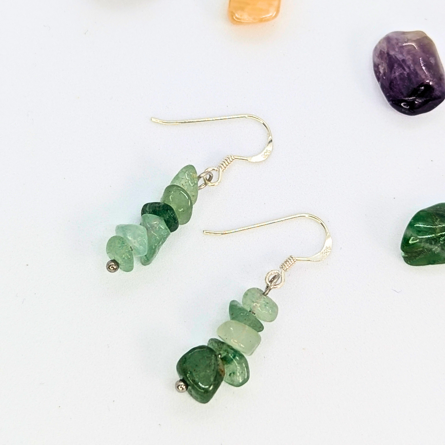Silver Sunbird Green Aventurine Earrings feature stacked green crystal chips on sterling silver dangle hooks, displayed on a white surface with colorful polished stones nearby.