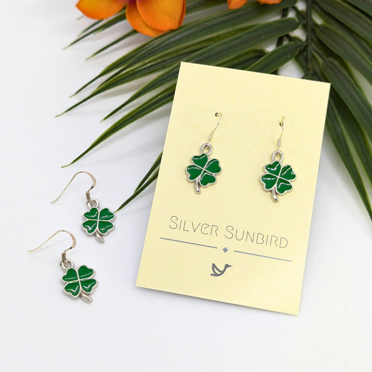 A pair of Lucky Four Leaf Clover Earrings by Silver Sunbird, featuring green charms, are displayed on a branded card. Another identical sterling silver pair is nearby with green leaves and orange flowers above them.