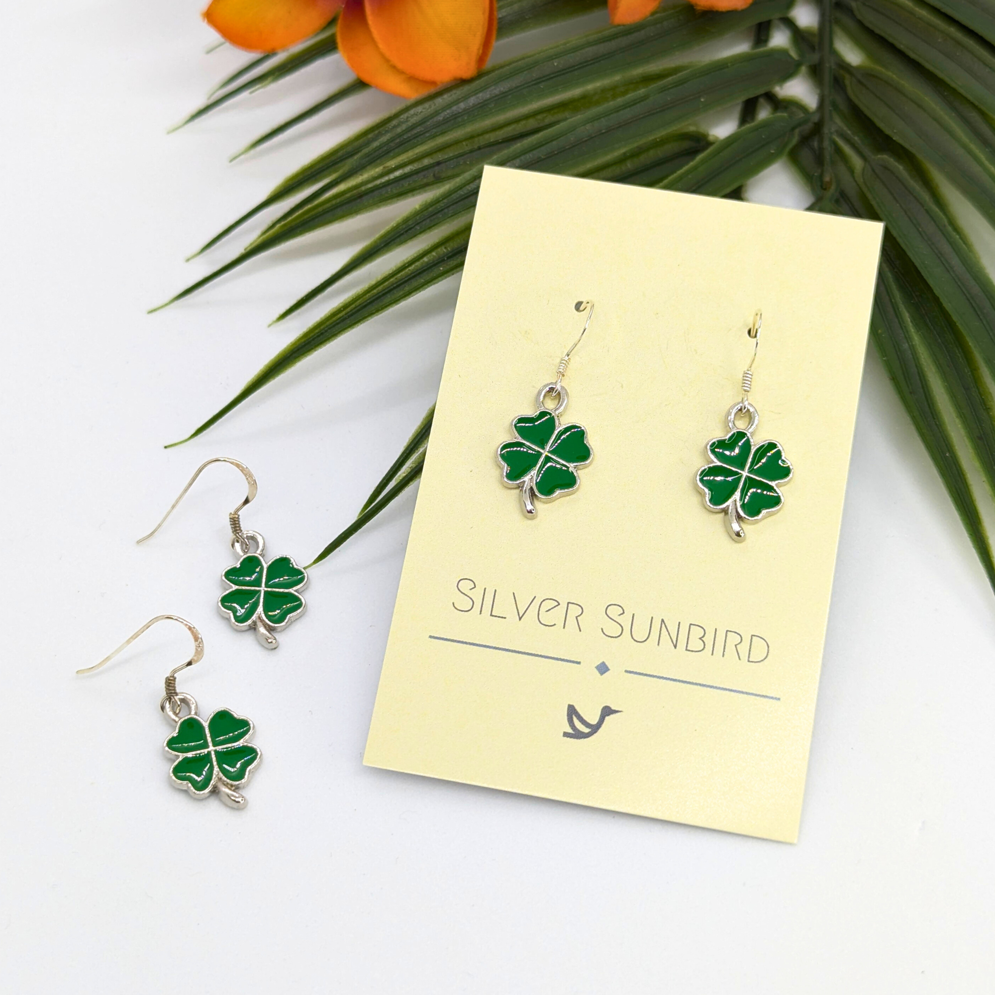 A pair of Lucky Four Leaf Clover Earrings by Silver Sunbird, featuring green charms, are displayed on a branded card. Another identical sterling silver pair is nearby with green leaves and orange flowers above them.