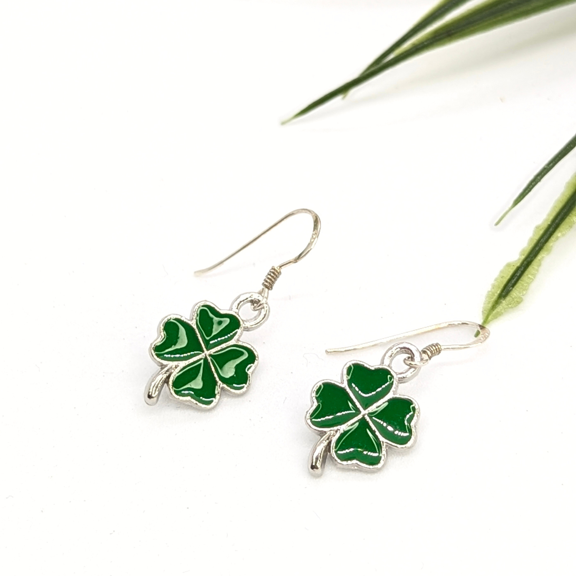 A pair of Silver Sunbird Lucky Four Leaf Clover Earrings with green enamel charms hangs from sterling silver hooks, displayed on a white background with green leaves in the corner.