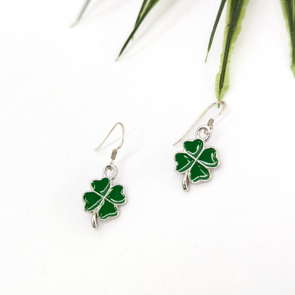 The Lucky Four Leaf Clover Earrings by Silver Sunbird, crafted in sterling silver with green enamel charms, are shown on a white surface with green leaves in the upper right corner.