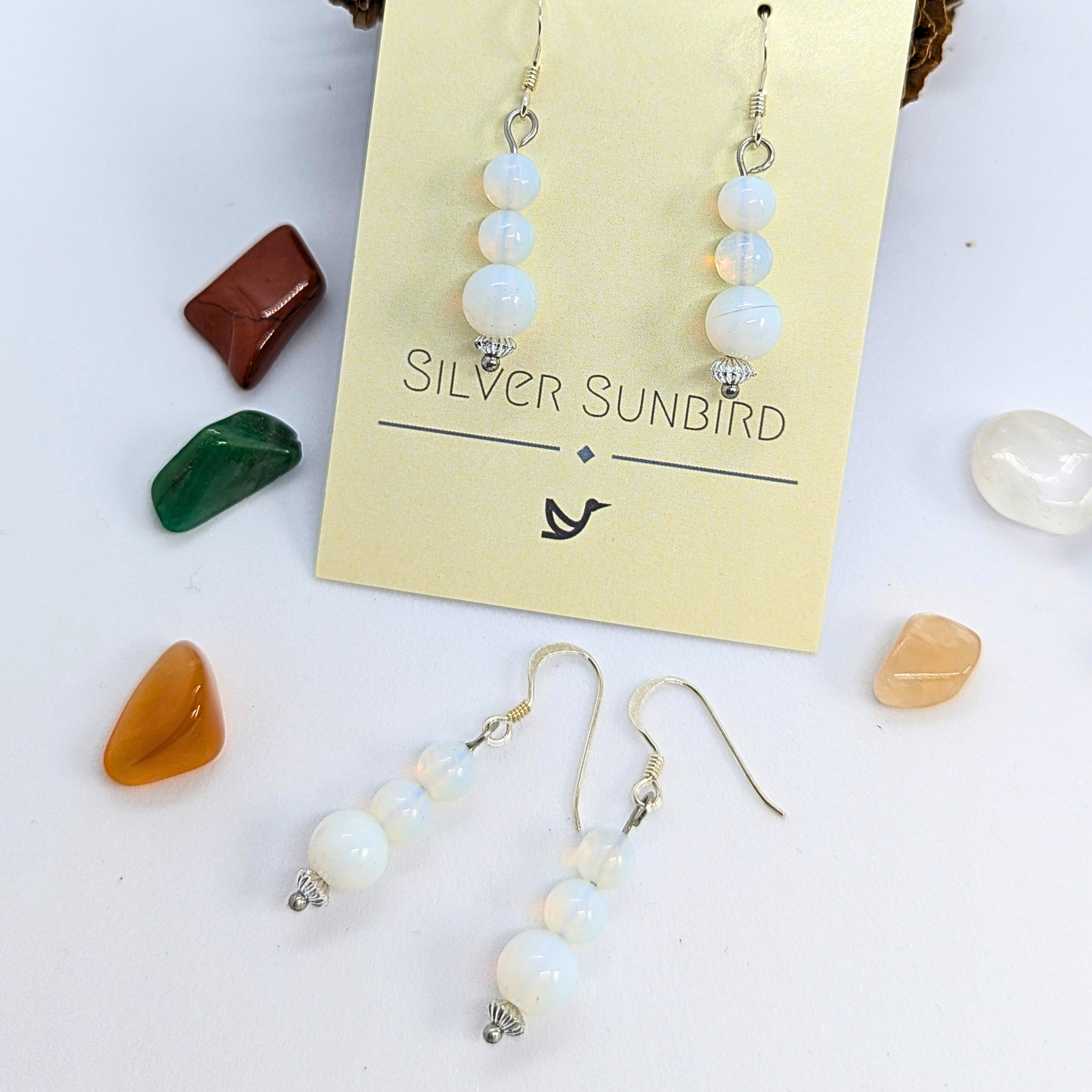 The Opal Dangle Earrings by Silver Sunbird, displayed on a branded card, feature round opalescent beads and are surrounded by polished stones in red, green, orange, and clear hues.