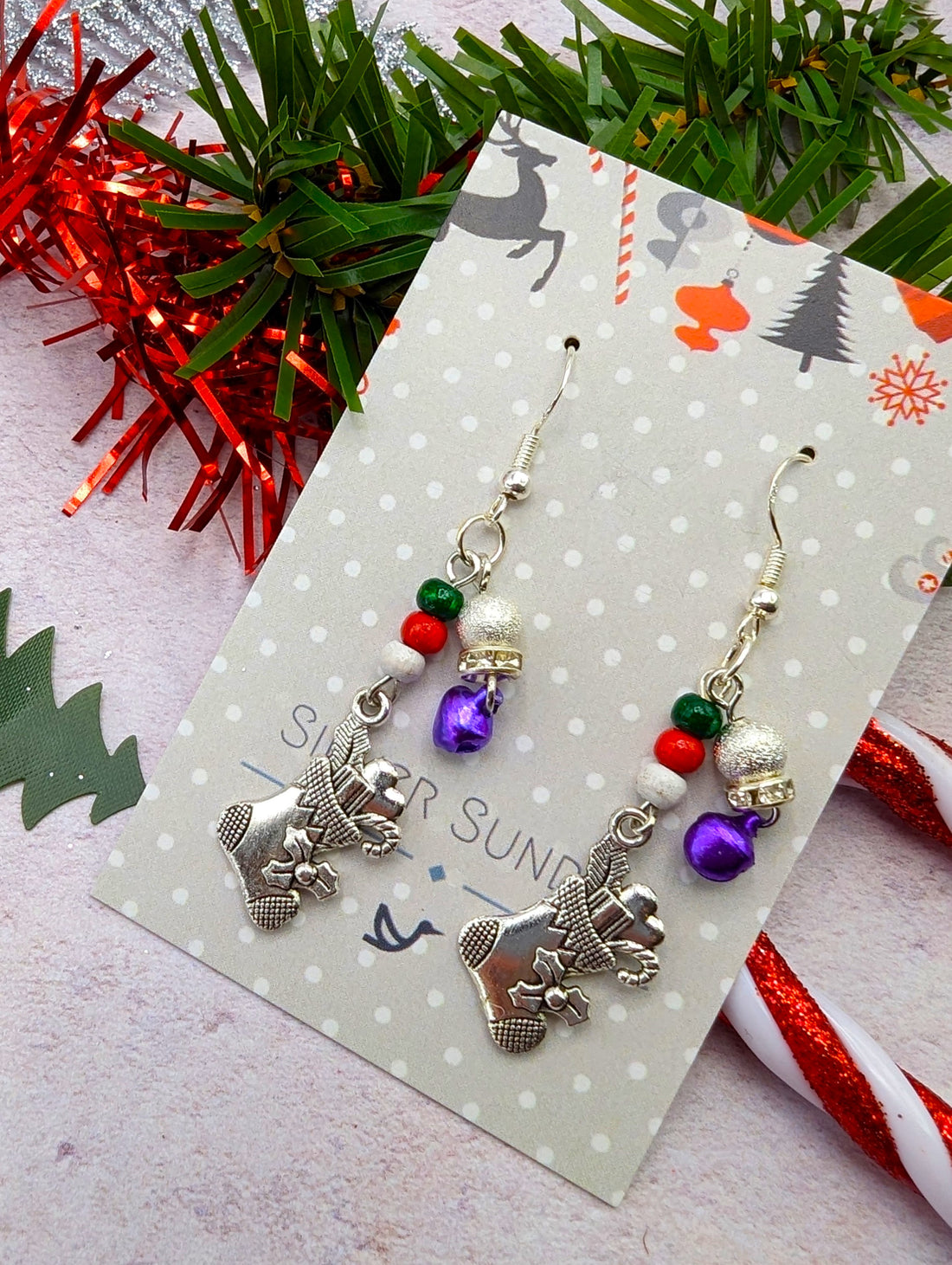 Introducing the Jingle Stockings Earrings by Silver Sunbird—festive Christmas earrings designed with sterling silver snowmen charms, purple bells, and red and green beads. They are beautifully displayed on a polka-dotted card with a background featuring red tinsel, greenery, and candy cane stripes. These earrings are perfect for adding a touch of handmade jewelry flair to your holiday ensemble.