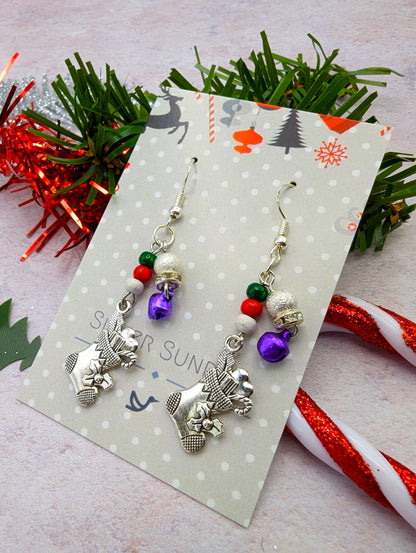 The Jingle Stockings Earrings by Silver Sunbird are festive and feature charming reindeer charms, red and green beads, and purple bells, all displayed on a decorative card. In the background, there&