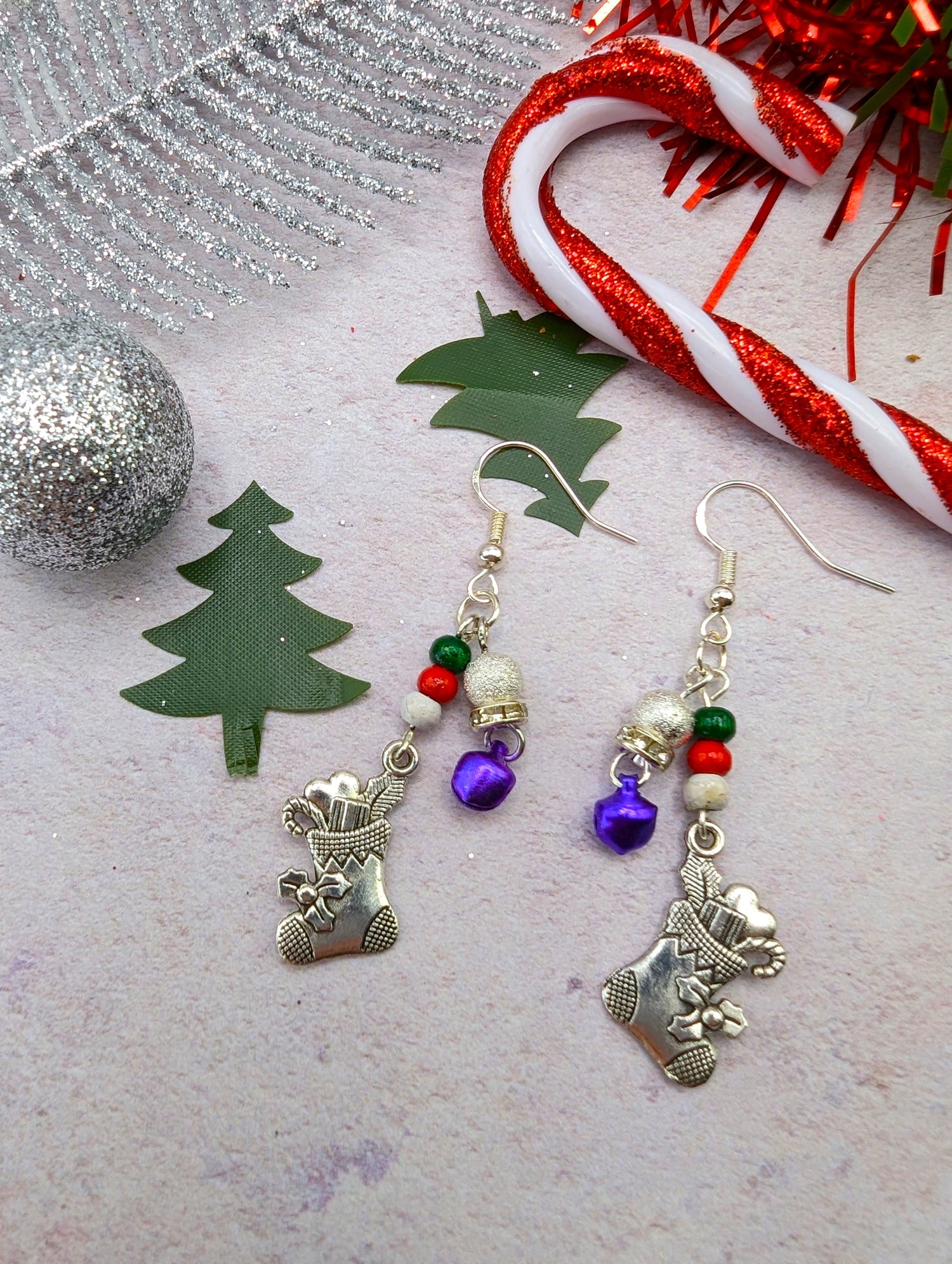 The Jingle Stockings Earrings by Silver Sunbird showcase festive Christmas-themed sterling silver stocking charms, adorned with small bells and beads in vibrant red, green, and purple. These earrings are artfully set against an arrangement of holiday decorations that include a silver ornament, green tree cutouts, and a candy cane ribbon on a light background.