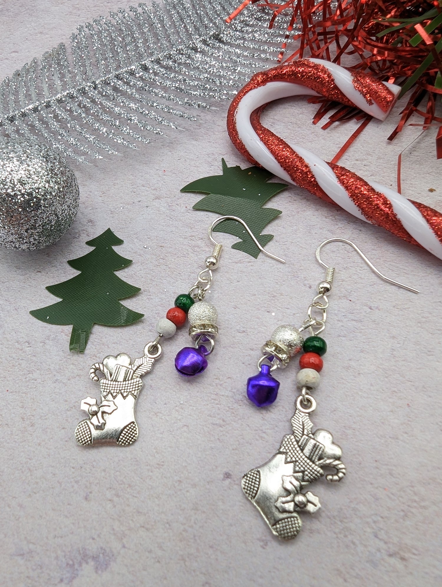 The Jingle Stockings Earrings by Silver Sunbird, showcasing handmade jewelry with silver stocking charms, purple bells, and beads, are presented on a surface decorated with sterling silver leaf, candy cane, silver bauble, and green tree shapes.