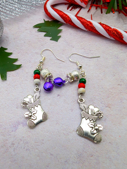 Introducing the Jingle Stockings Earrings from Silver Sunbird—festive Christmas earrings featuring silver stockings and vibrant beads in red, green, and purple. This handmade jewelry piece is beautifully adorned with a cane ornament on a light background, accented by red and white candy canes and green holly leaves.