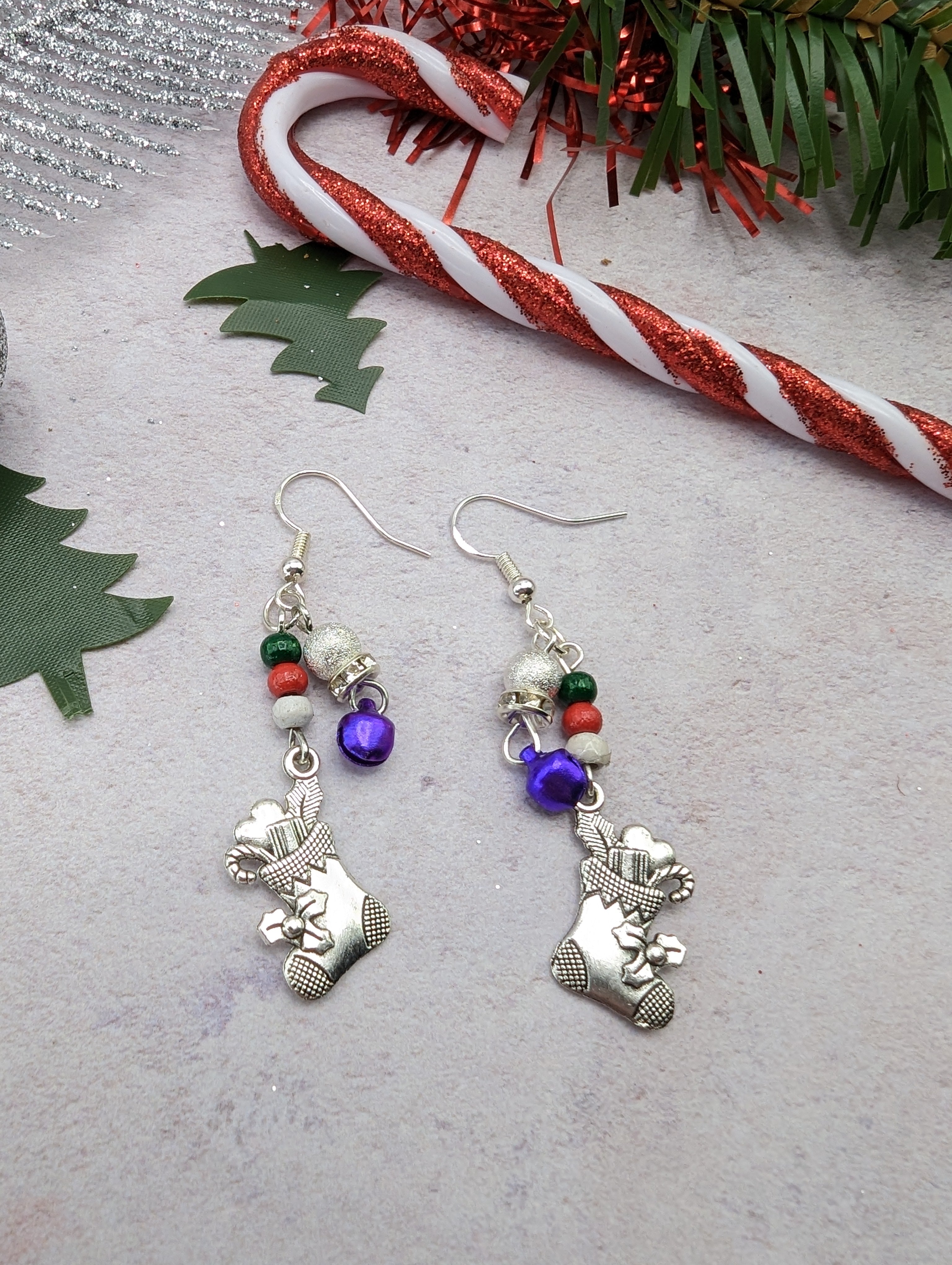 The Jingle Stockings Earrings by Silver Sunbird are showcased on a light surface, featuring sterling silver teddy bears, red and green beads, and small purple bells. Nearby sprigs of green foliage and a glittery candy cane decoration complement the handmade jewelry&