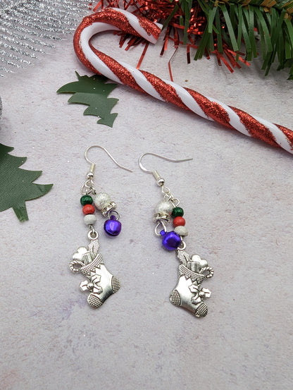 The Jingle Stockings Earrings by Silver Sunbird are showcased on a light surface, featuring sterling silver teddy bears, red and green beads, and small purple bells. Nearby sprigs of green foliage and a glittery candy cane decoration complement the handmade jewelry&