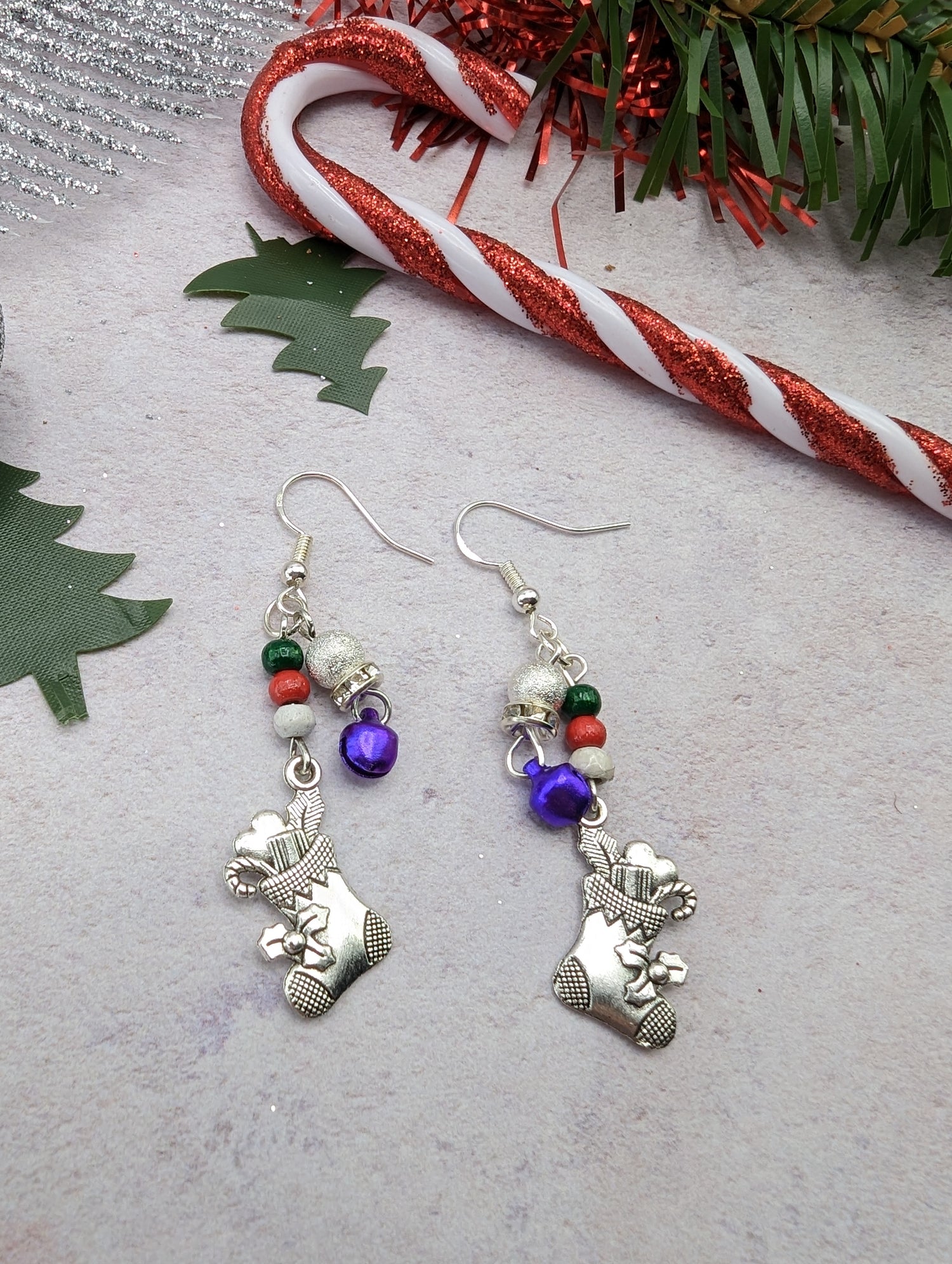 The Jingle Stockings Earrings by Silver Sunbird are festive Christmas earrings featuring sterling silver charms of Christmas stockings, adorned with red, green, and pearl beads. A candy cane and green tree decorations rest on a light surface in the background, perfectly capturing the essence of handmade jewelry.