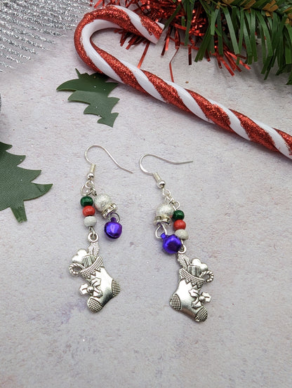 The Jingle Stockings Earrings by Silver Sunbird are festive Christmas earrings featuring sterling silver charms of Christmas stockings, adorned with red, green, and pearl beads. A candy cane and green tree decorations rest on a light surface in the background, perfectly capturing the essence of handmade jewelry.
