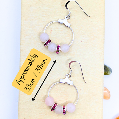 A pair of Rose Quartz Statement Earrings from Silver Sunbird, featuring pink beads and small red stones on the bottom half, is displayed on a beige background. Each hypoallergenic, circular earring measures approximately 3.9 cm (39 mm) in length, as indicated by a yellow label with black text and arrows pointing to the earrings.