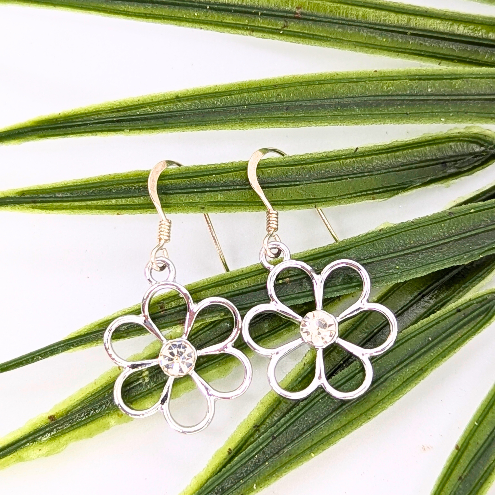 Sparkly Silver Daisy Earrings by Silver Sunbird, featuring clear gemstone centers, are displayed on green palm leaves against a white background.