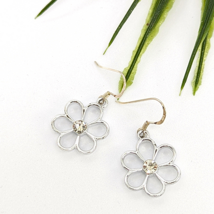 The Sparkly Silver Daisy Earrings by Silver Sunbird feature sterling silver floral designs with clear gemstone centers, shown on a white surface accented by green leaves.