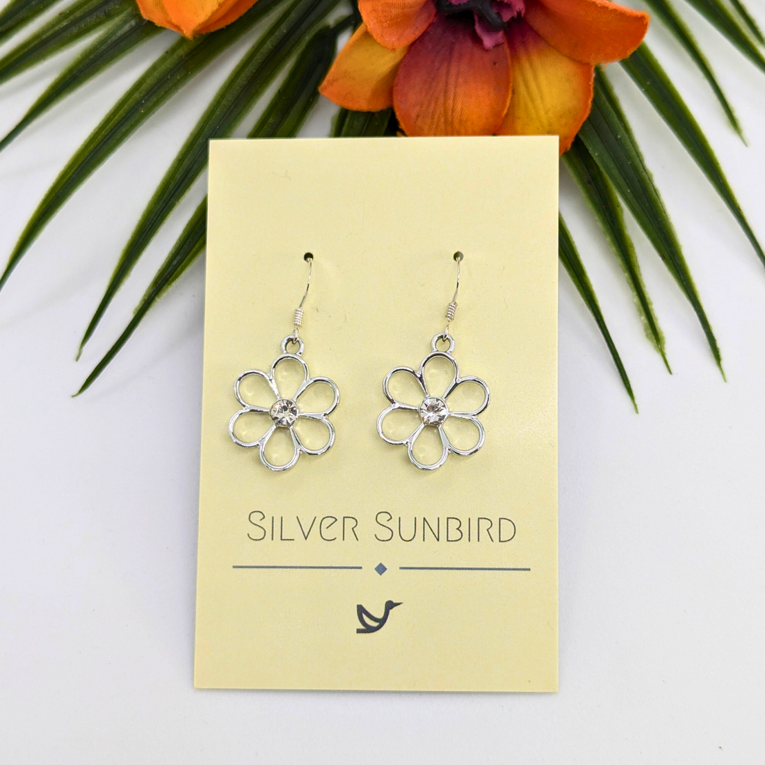 Sparkly Silver Daisy Earrings by Silver Sunbird, crafted in sterling silver with a central gem, are displayed on a cream &quot;SILVER SUNBIRD&quot; card among green leaves and orange flowers for an elegant floral effect.