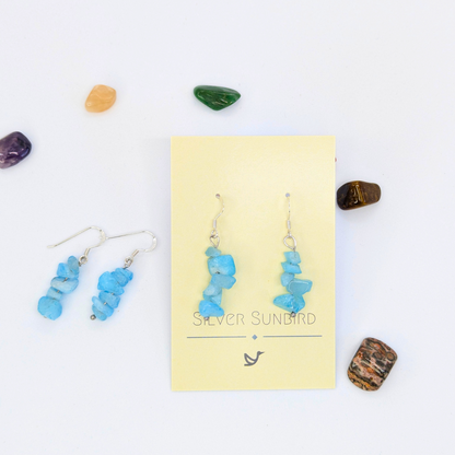 Aquamarine Gemstone Earrings by Silver Sunbird displayed on a yellow branded card, surrounded by colorful stones. A matching pair of sterling silver hypoallergenic earrings rests to the left on a white background.